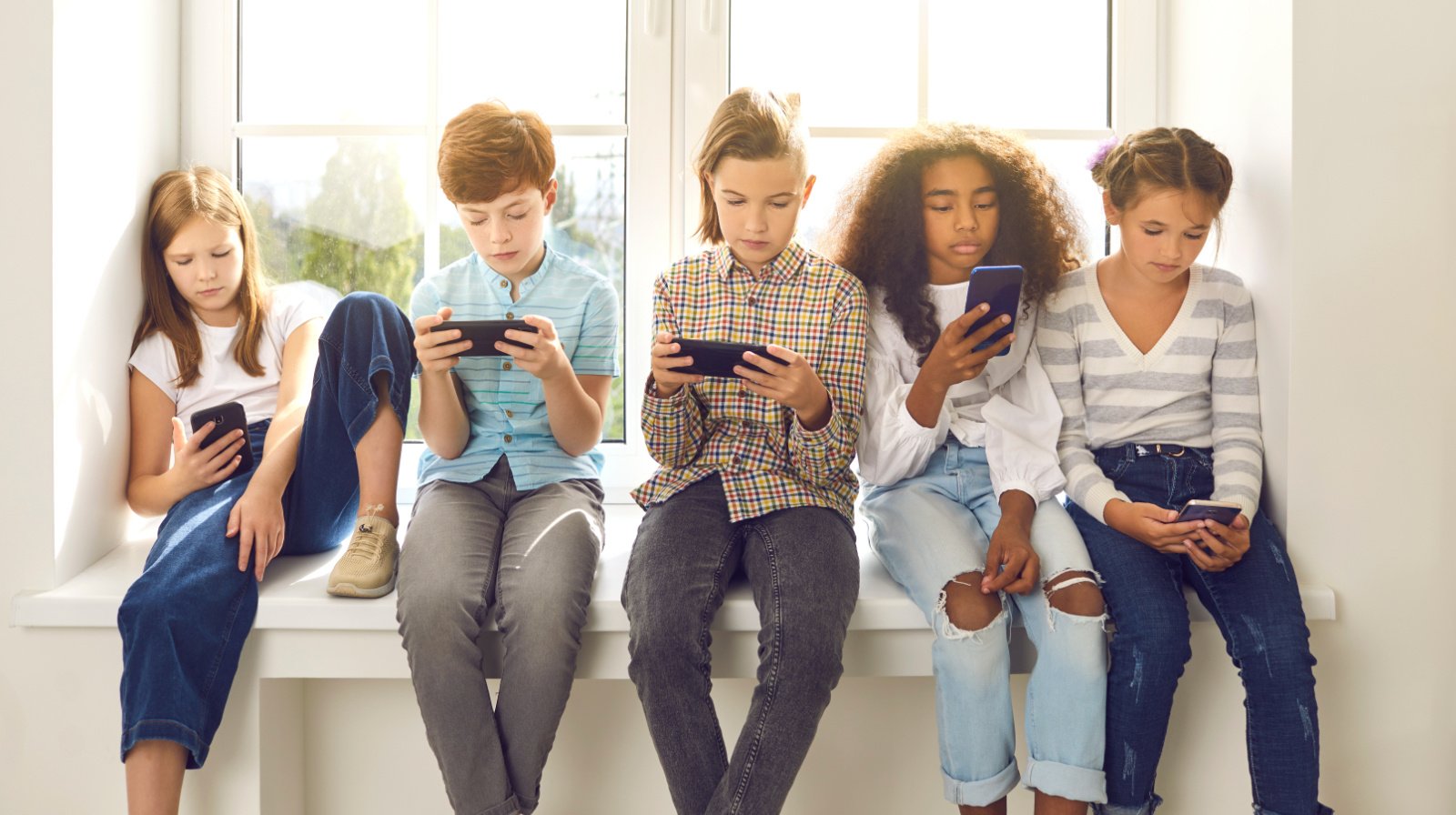 A group of preteens sitting on a window sill while using their phones.